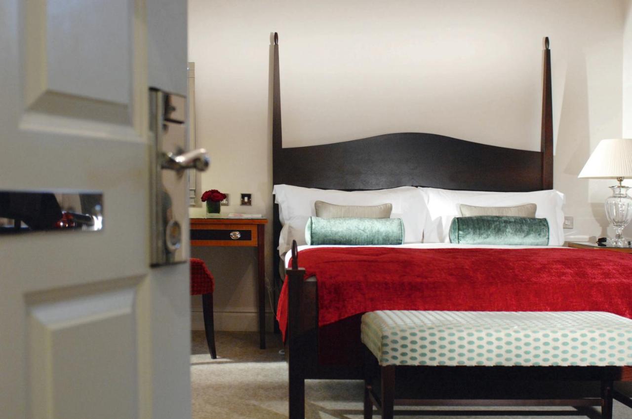 Macdonald Bath Spa Hotel Δωμάτιο φωτογραφία The photo shows a well-decorated hotel room. In the foreground, there is a partially opened door leading into the room. The focal point of the room is a large bed with a dark wooden headboard and stylish red and white bedding, accented with green pil