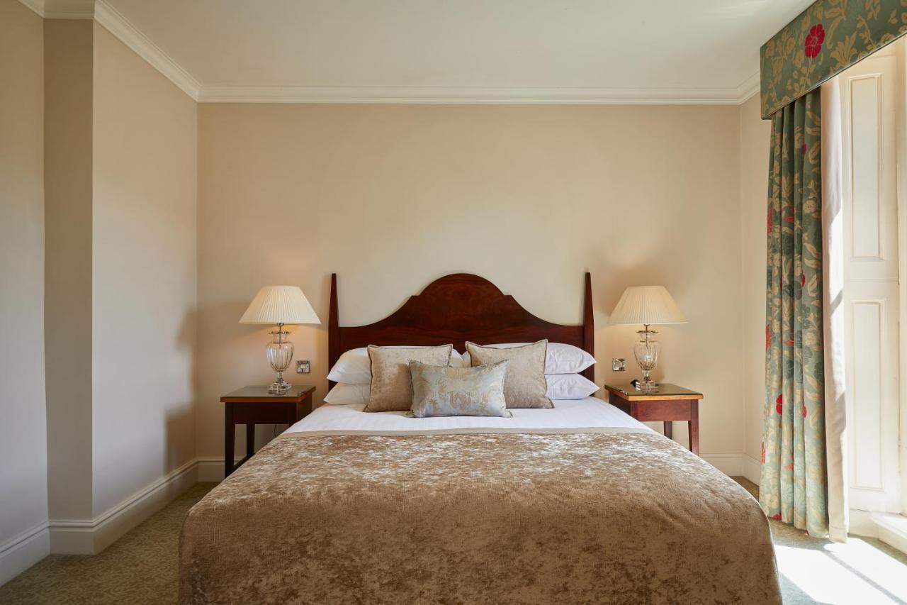 Macdonald Bath Spa Hotel Εξωτερικό φωτογραφία The photo shows a cozy hotel bedroom. It features a large bed with a plush, light-colored bedspread and several pillows. On either side of the bed, there are wooden nightstands with lamps providing soft lighting. The walls are painted a neutral color