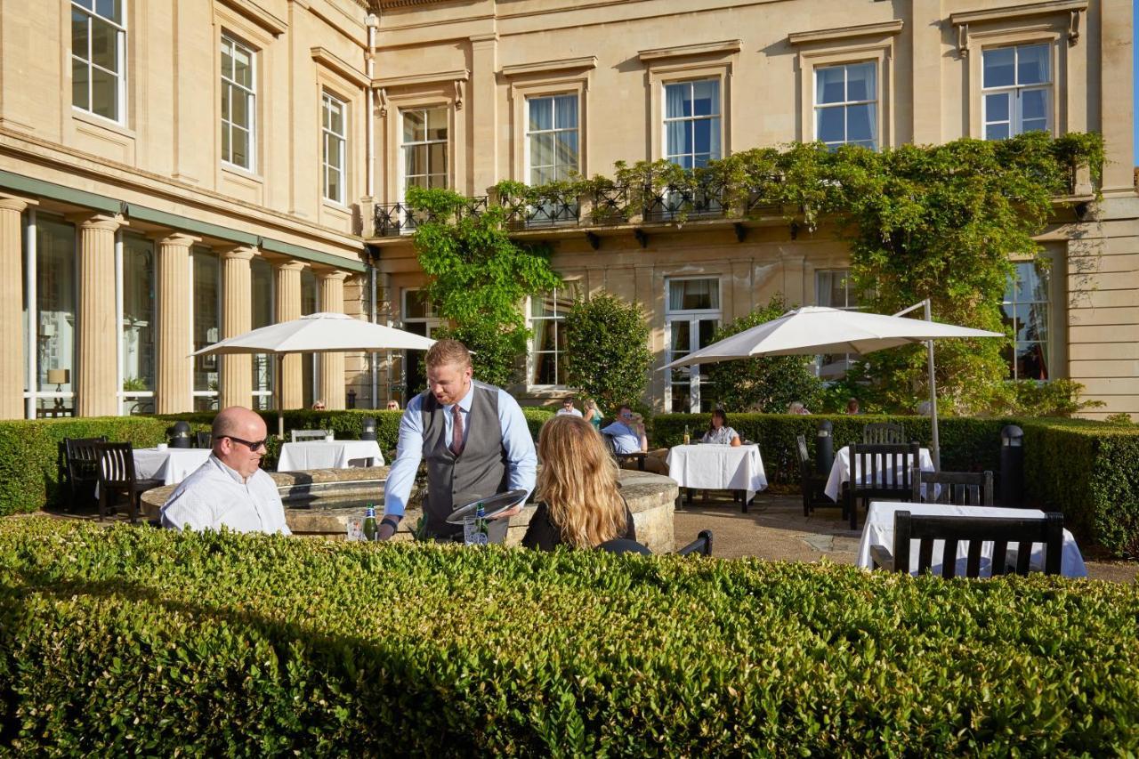 Macdonald Bath Spa Hotel Εξωτερικό φωτογραφία The photo depicts an outdoor dining area of a restaurant. In the foreground, there are neatly trimmed hedges surrounding the dining space. Two patrons are seated at a table; one is a man wearing sunglasses and the other is a woman facing him. A waite