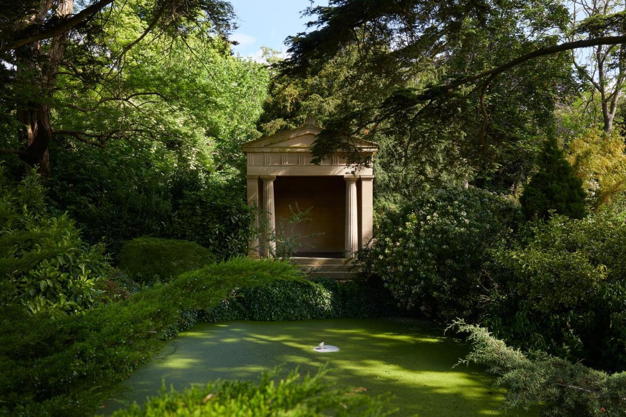 Macdonald Bath Spa Hotel Εξωτερικό φωτογραφία The photo shows a serene garden scene featuring lush greenery and a tranquil pond. In the background, there is a classical-looking structure with columns, resembling a small temple or pavilion. The pond appears to have vegetation covering its surface