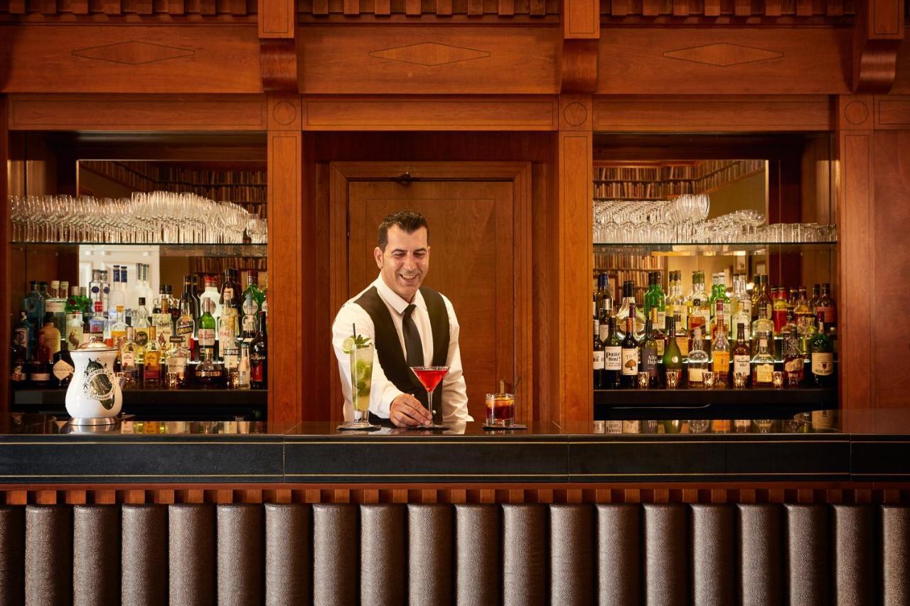 Macdonald Bath Spa Hotel Εξωτερικό φωτογραφία The photo depicts a bartender standing behind a polished bar. He is wearing a formal outfit with a vest and a bow tie, and he appears to be smiling as he presents a cocktail. Behind him is a well-stocked bar shelf filled with various bottles of liquo