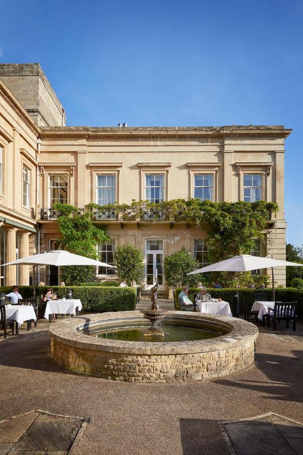 Macdonald Bath Spa Hotel Εξωτερικό φωτογραφία The photo depicts a charming outdoor dining area of a classic-looking building with elegant architecture. The structure features large windows and is adorned with greenery, including climbing plants. In the foreground, there is a circular fountain, a