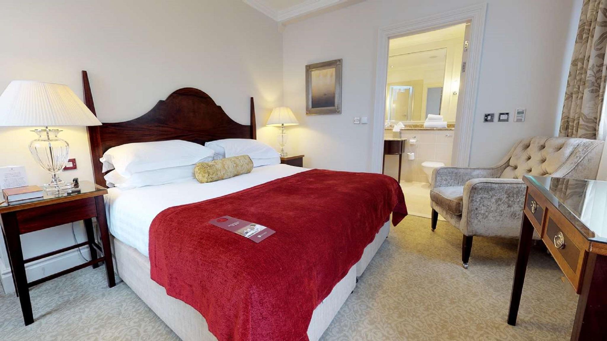 Macdonald Bath Spa Hotel Εξωτερικό φωτογραφία The photo shows a well-furnished hotel room. In the center, there is a large bed with white linens and a red throw blanket. A decorative pillow is placed on the bed. On either side of the bed, there are bedside tables with lamps. To the right, there 