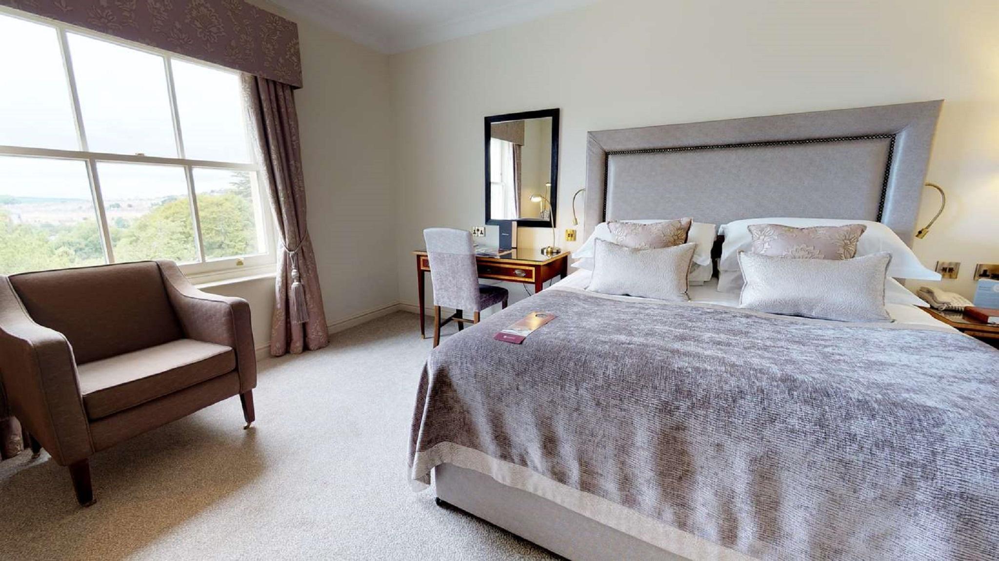 Macdonald Bath Spa Hotel Εξωτερικό φωτογραφία The photo shows a cozy and well-decorated bedroom. It features a large bed with a plush, grey bedspread and several decorative pillows. Beside the bed, there is a wooden bedside table with a lamp. A small desk and chair are positioned near a window, 