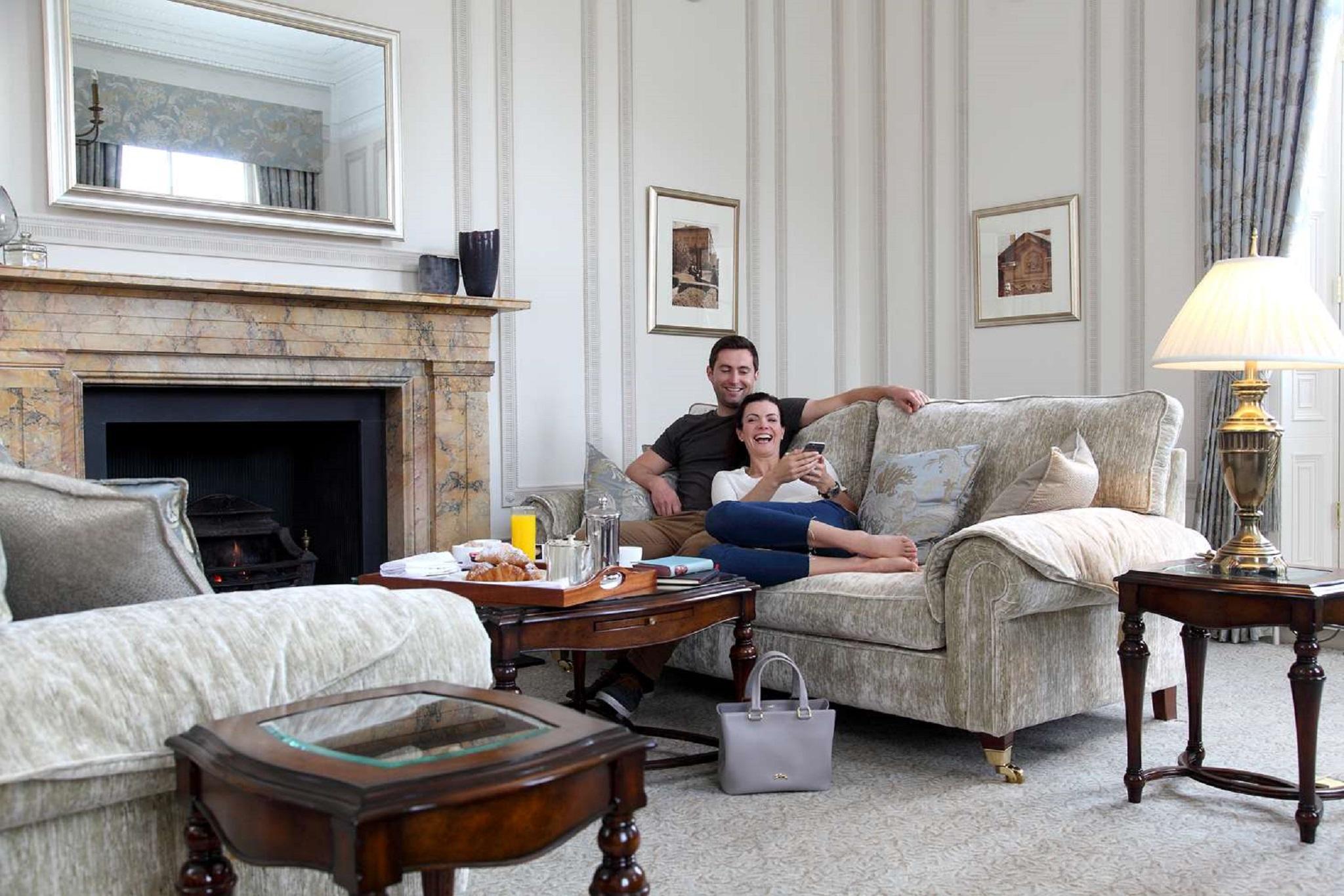 Macdonald Bath Spa Hotel Εξωτερικό φωτογραφία The photo shows a cozy living room setting where a couple is sitting together on a light-colored sofa. They appear relaxed, with the man sitting slightly behind the woman, who is smiling and looking at her phone. The room features elegant decor, incl