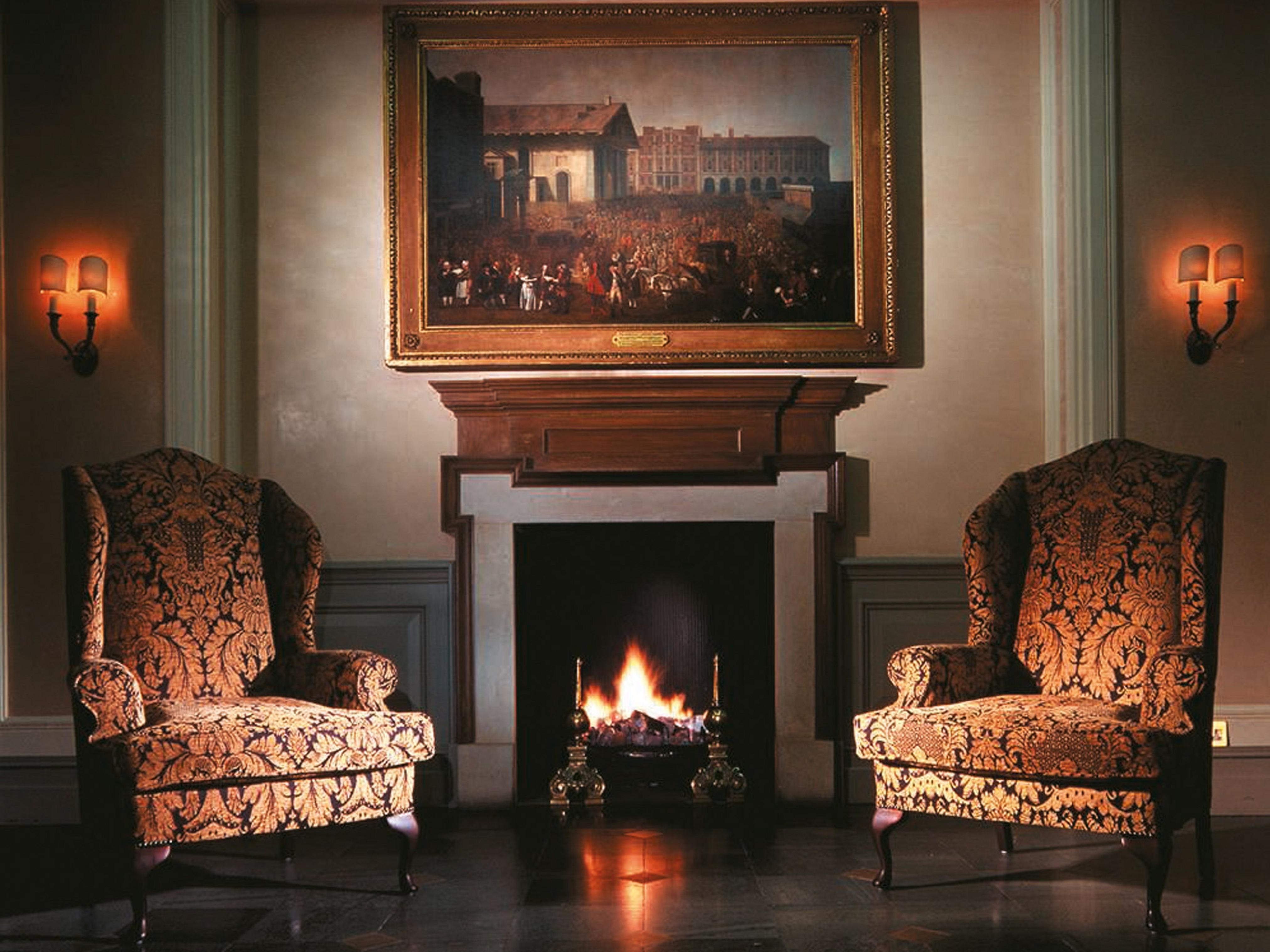 Macdonald Bath Spa Hotel Εξωτερικό φωτογραφία The image shows a cozy indoor setting featuring two ornate armchairs placed opposite a fireplace. Above the fireplace hangs a large painting in a decorative frame. The warm glow from the fire illuminates the room, and there are wall sconces providing