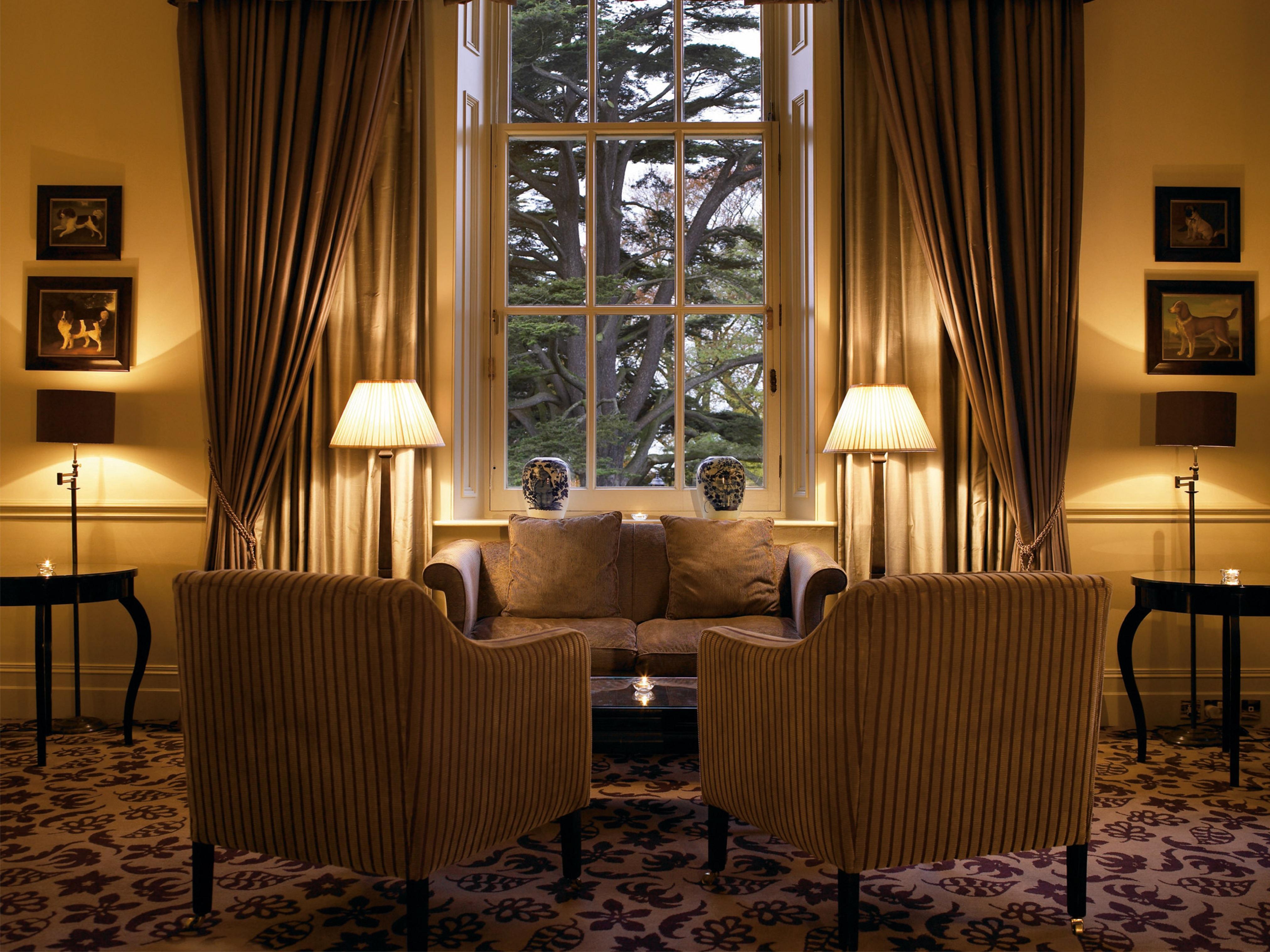 Macdonald Bath Spa Hotel Εξωτερικό φωτογραφία The photo shows a cozy and elegant sitting area. There are two upholstered armchairs facing a small sofa, all arranged around a central coffee table. Soft lighting is provided by two lamps with white shades, one beside each chair. The window behind t