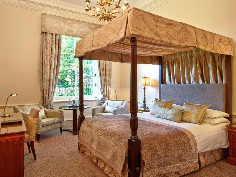Macdonald Bath Spa Hotel Δωμάτιο φωτογραφία The photo shows an elegantly designed bedroom featuring a four-poster bed with a decorative canopy. The bed is dressed with luxurious bedding and pillows, creating a cozy and inviting atmosphere. Flanking the bed are two comfortable armchairs and a s