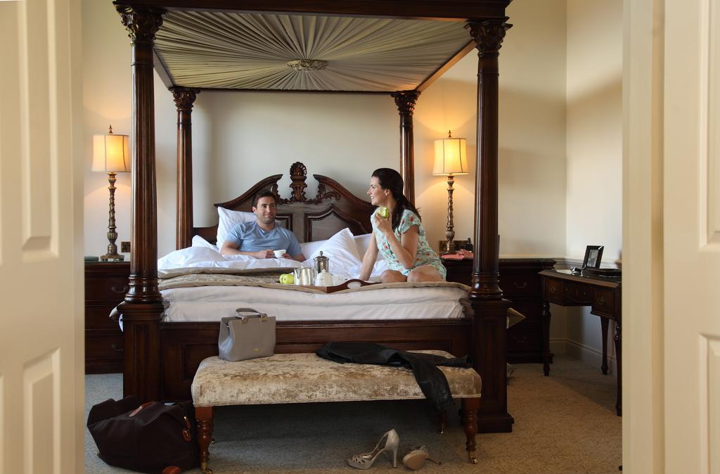 Macdonald Bath Spa Hotel Εξωτερικό φωτογραφία The image depicts a spacious, well-lit bedroom featuring a large four-poster bed with an ornate headboard. A man and a woman are sitting on the bed, appearing to engage in conversation. The woman is dressed in a patterned outfit, while the man is wea