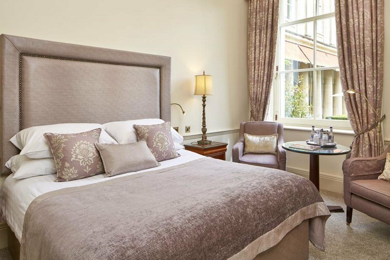 Macdonald Bath Spa Hotel Εξωτερικό φωτογραφία The photo shows a stylish and elegantly decorated hotel room. It features a large bed with soft linens and several decorative pillows. The bedspread is a muted shade of beige or taupe. Beside the bed, there's a small table with two armchairs. One of 
